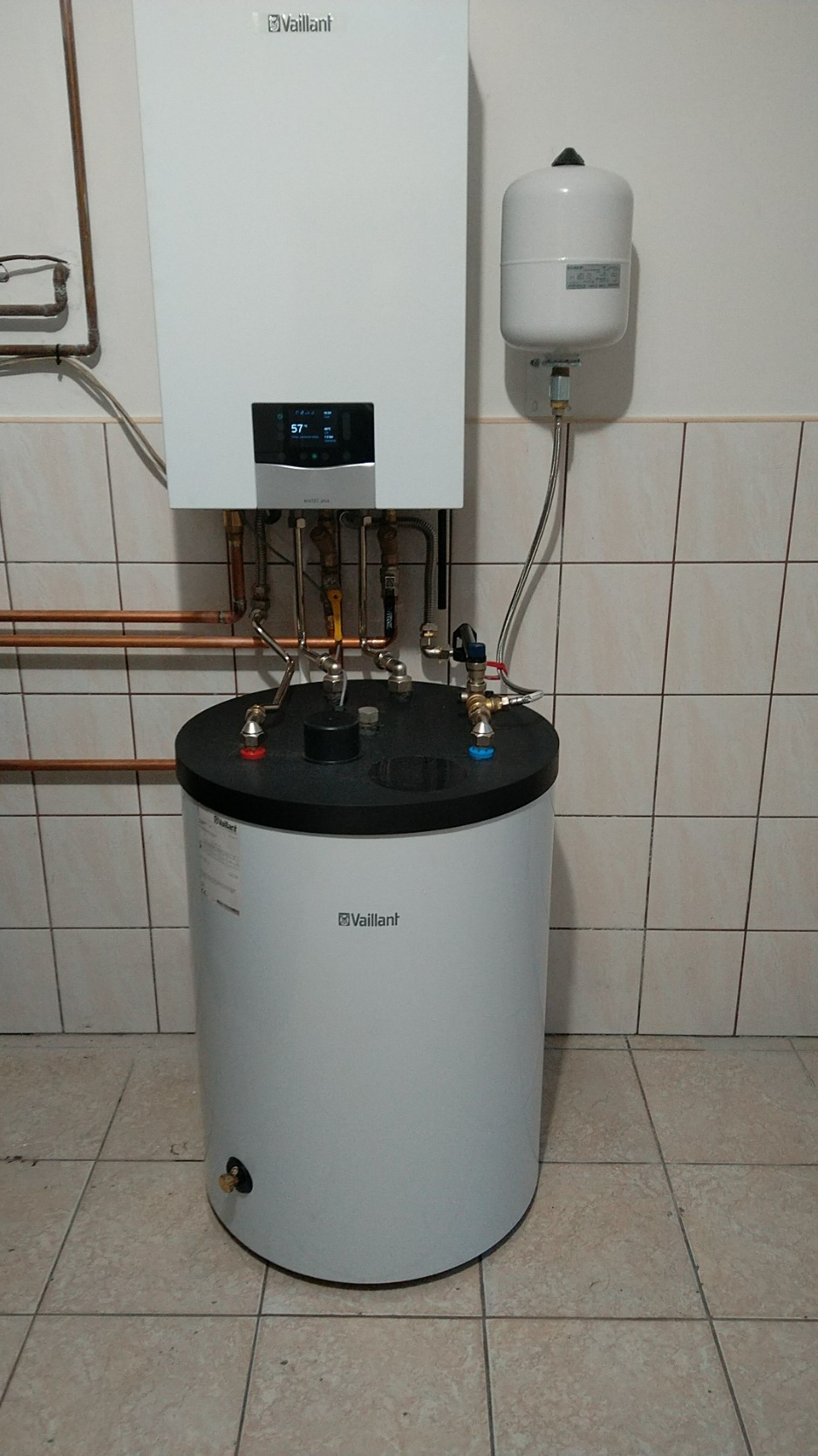 Biały piec gazowy Vaillant z podgrzewaczem wody, zamontowany na tle kafelkowanej ściany i podłogi, widoczne miedziane rury i zbiornik wyrównawczy.