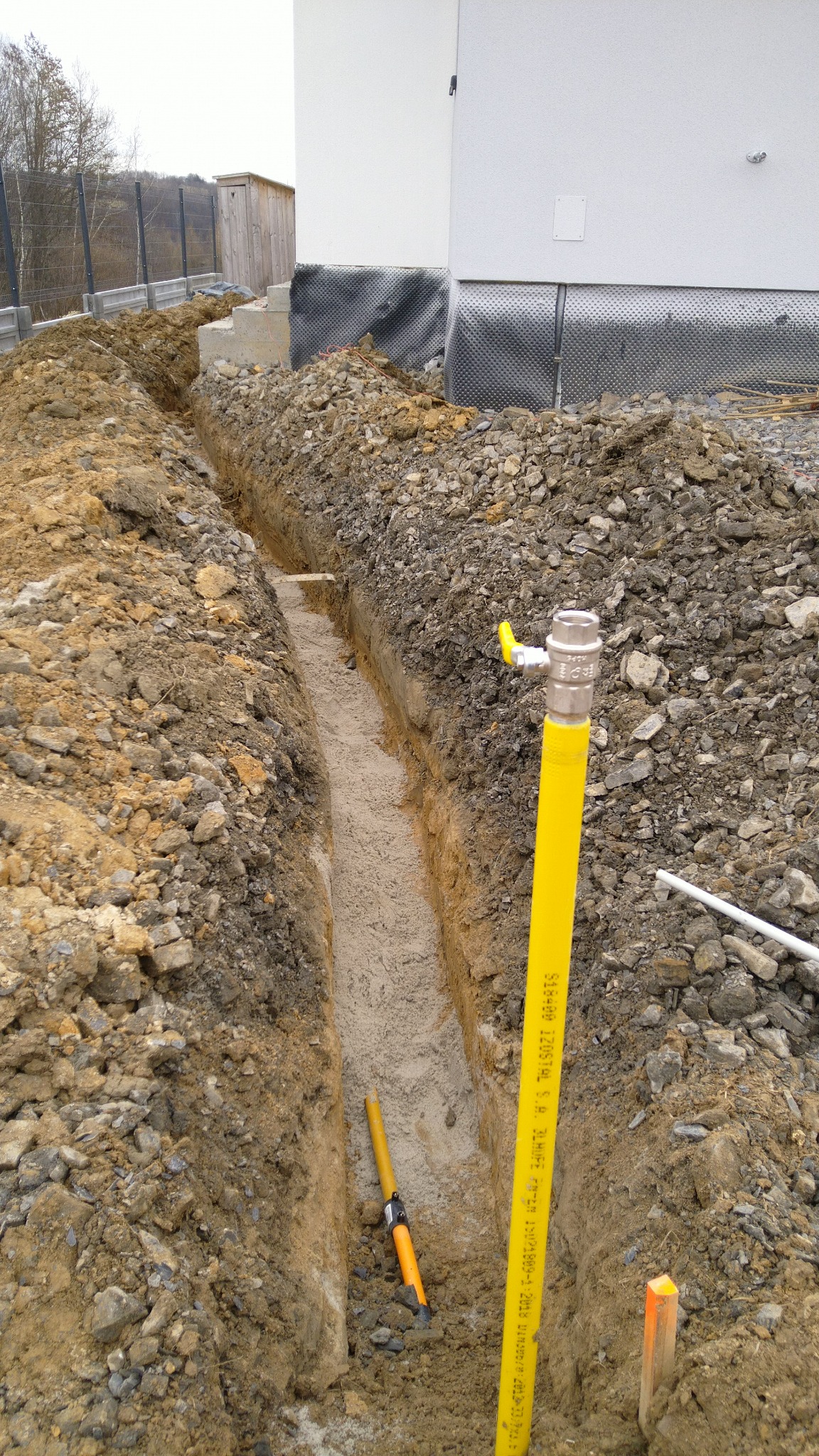 Wykopany wąski rów w ziemi z ułożonym piaskowym podłożem, widoczna żółta rura gazowa z zaworem oraz fragment instalacji gazowej w wykopie na tle fundamentów budynku.