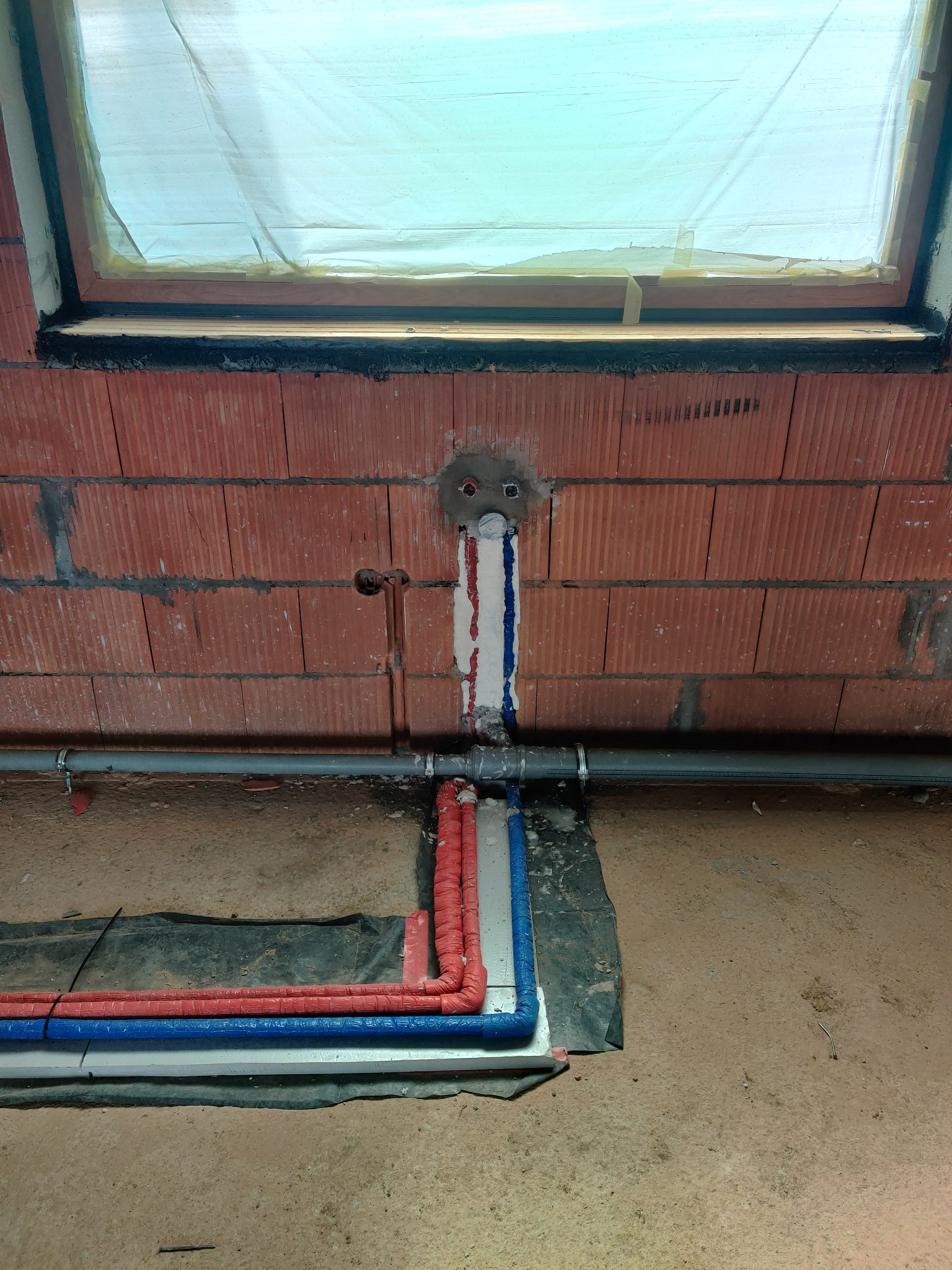 Surowy stan instalacji wodnej i grzewczej w pomieszczeniu z oknem dachowym: widoczne rury w izolacji czerwonej i niebieskiej prowadzone po podłodze, oraz pionowe podejścia do instalacji w ścianie...