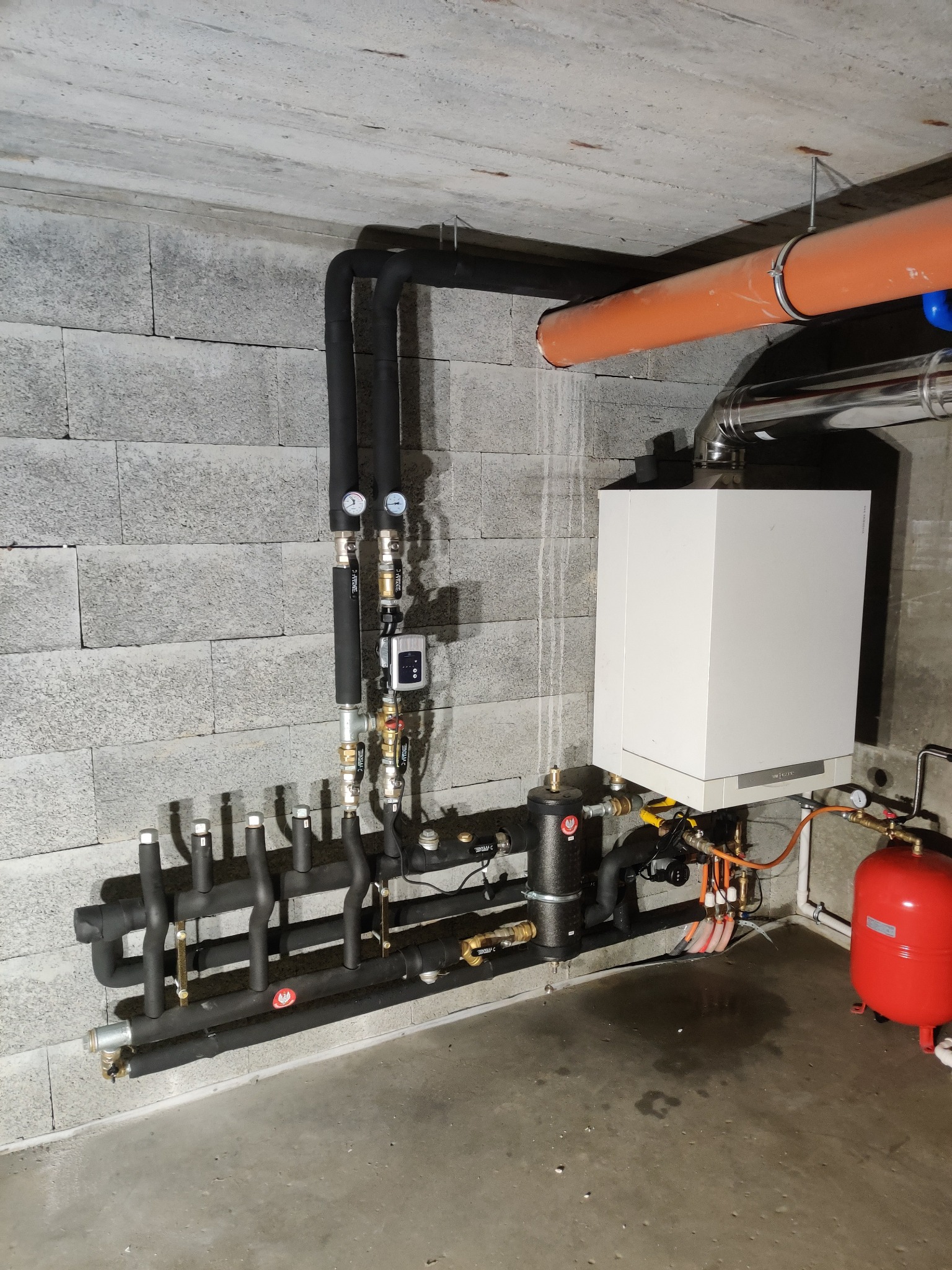 Nowoczesny piec grzewczy Viessmann zamontowany w surowym wnętrzu, z widocznymi rurami w izolacji i czerwonym zbiornikiem wyrównawczym, na tle ściany z szarego betonu komórkowego.