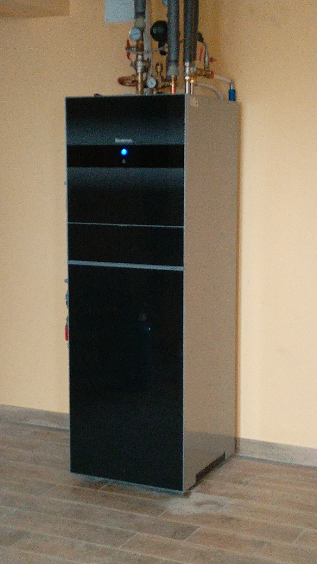 Nowoczesny piec gazowy z czarnym frontem i srebrnymi bokami, zainstalowany przy beżowej ścianie na podłodze z paneli imitujących drewno, widoczne rury i zawory instalacji co.