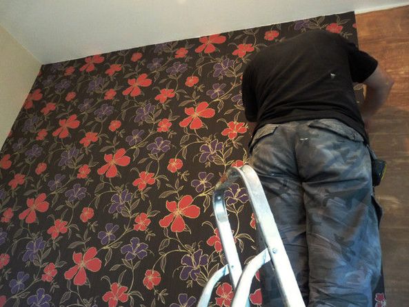 Pracownik na drabinie aplikuje wzorzystą tapetę w kwiaty na ścianę, widoczne narzędzia w kieszeni spodni roboczych.