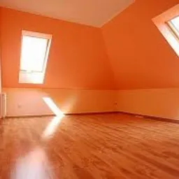 Poddasze po odświeżeniu: pomarańczowe ściany, jasne okna dachowe w skosach, parkiet na podłodze z widocznymi promieniami słońca, biały pas u dołu ściany.