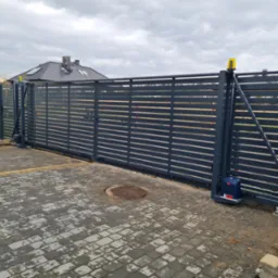 Szara, przesuwna brama wjazdowa z poziomymi panelami, wyposażona w niebieski siłownik automatyczny i żółtą lampę ostrzegawczą, zainstalowana na posesji z kostką brukową i widocznym włazem...