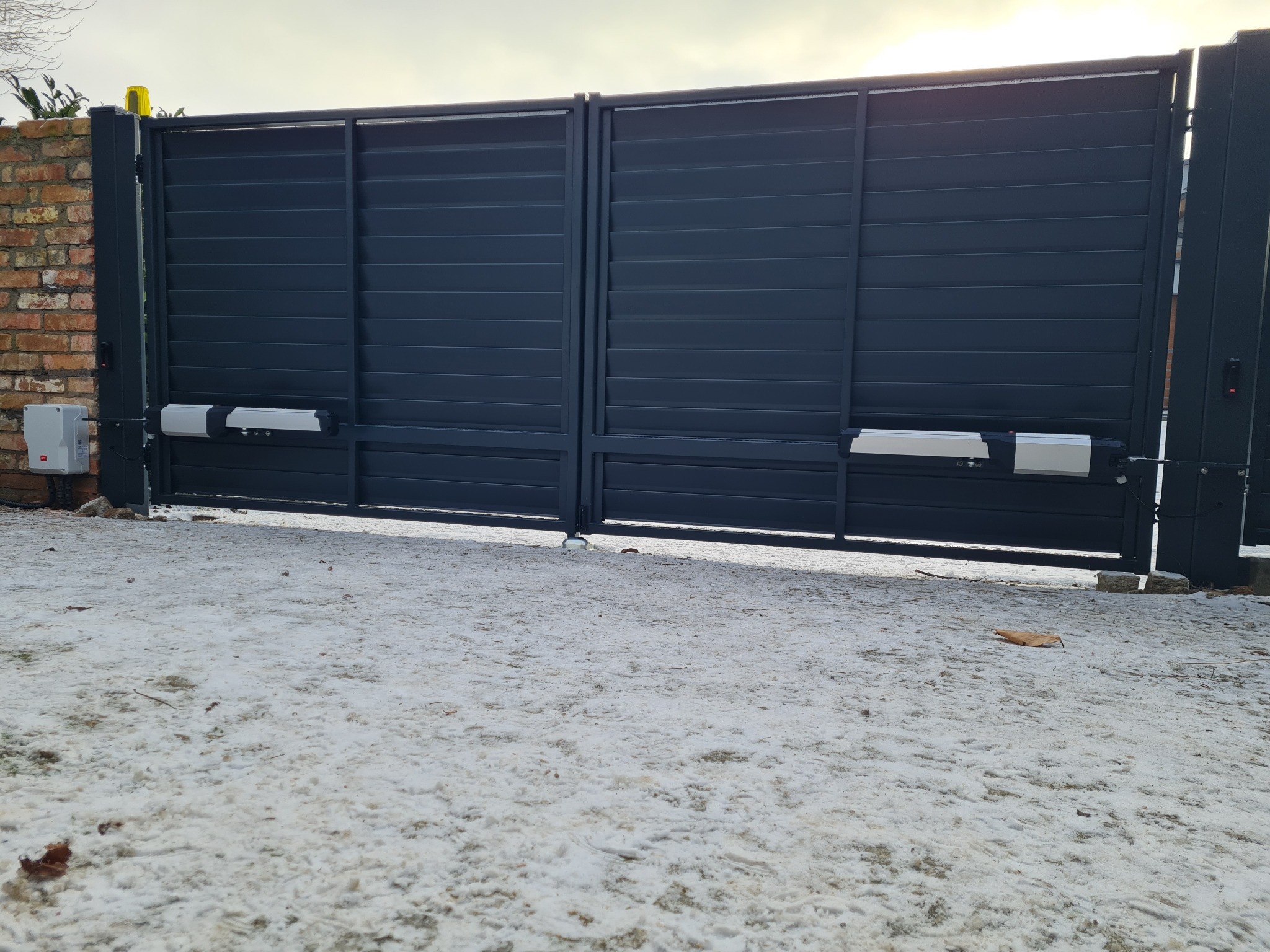 Nowoczesna, przesuwna brama wjazdowa w kolorze antracytowym z zamontowaną automatyką, widoczna na tle ośnieżonego podjazdu i ceglanego muru.
