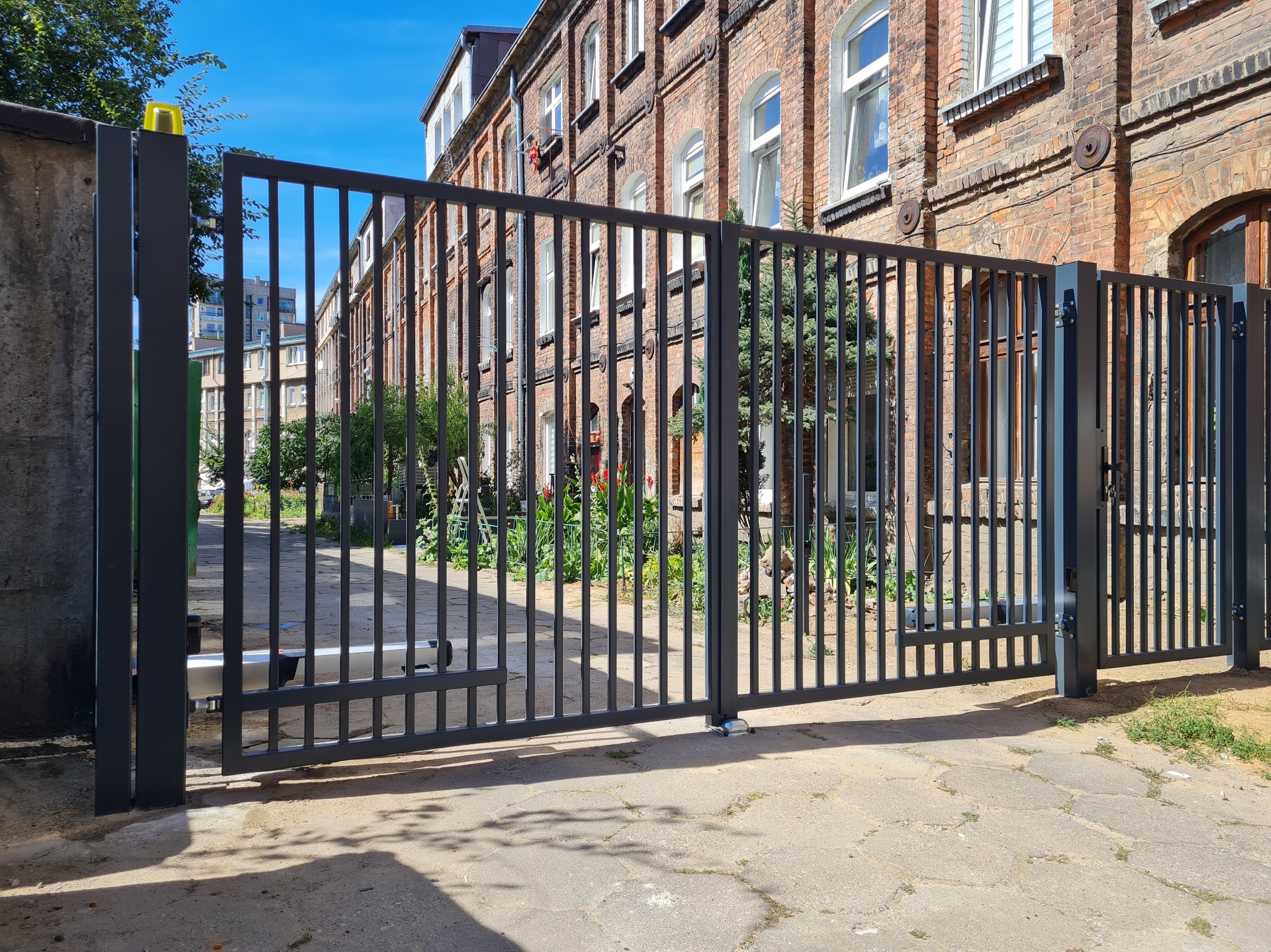 Nowoczesna, automatyczna brama wjazdowa z pionowymi szczeblami, częściowo otwarta, z żółtą lampą ostrzegawczą, na tle ceglanego budynku osiedla.