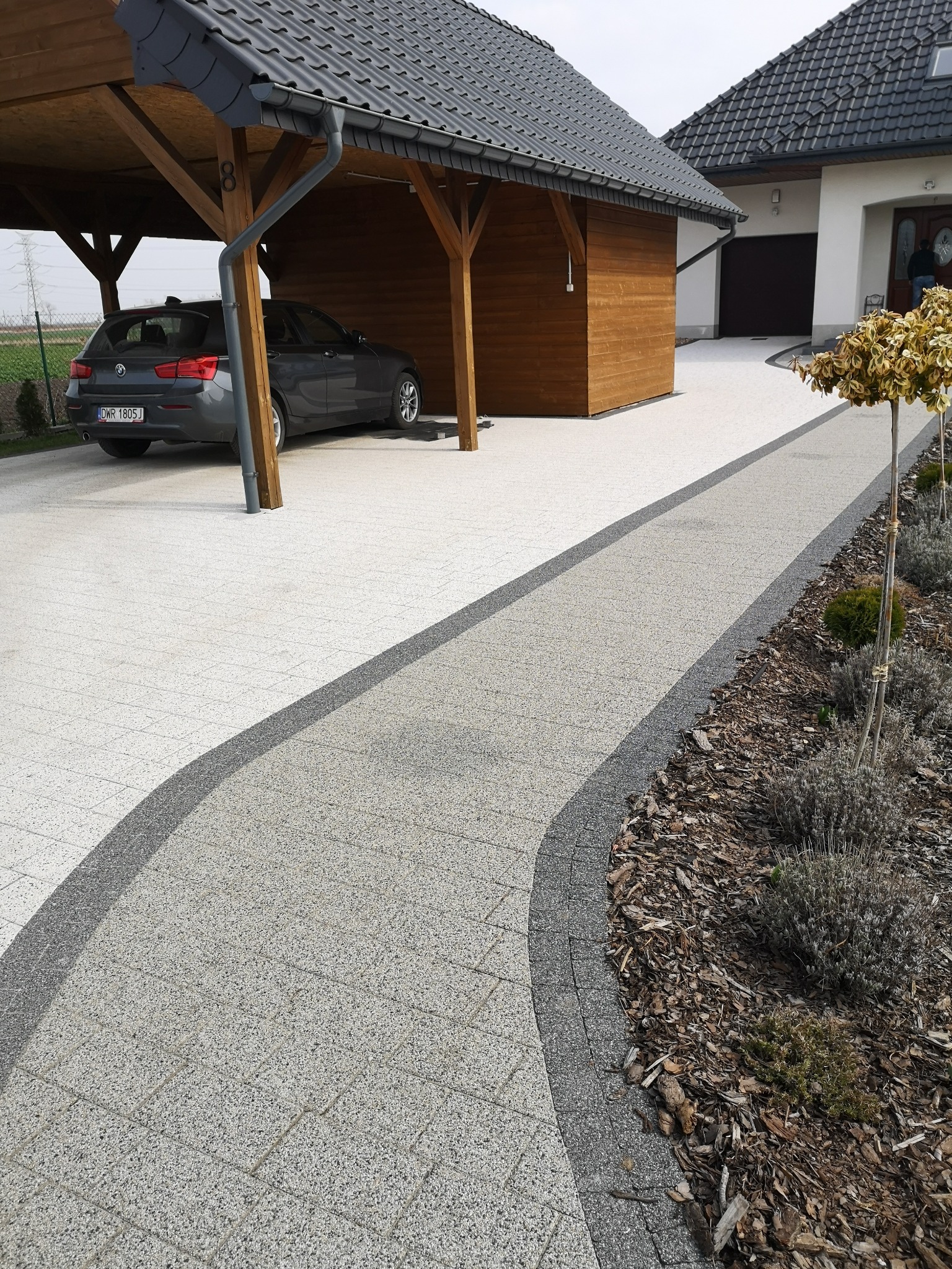 Jasnoszara nawierzchnia z kostki brukowej z ciemnoszarym obramowaniem, prowadząca od wiaty samochodowej do domu, z rabatą obsypaną korą i ozdobnymi drzewkami po prawej stronie.