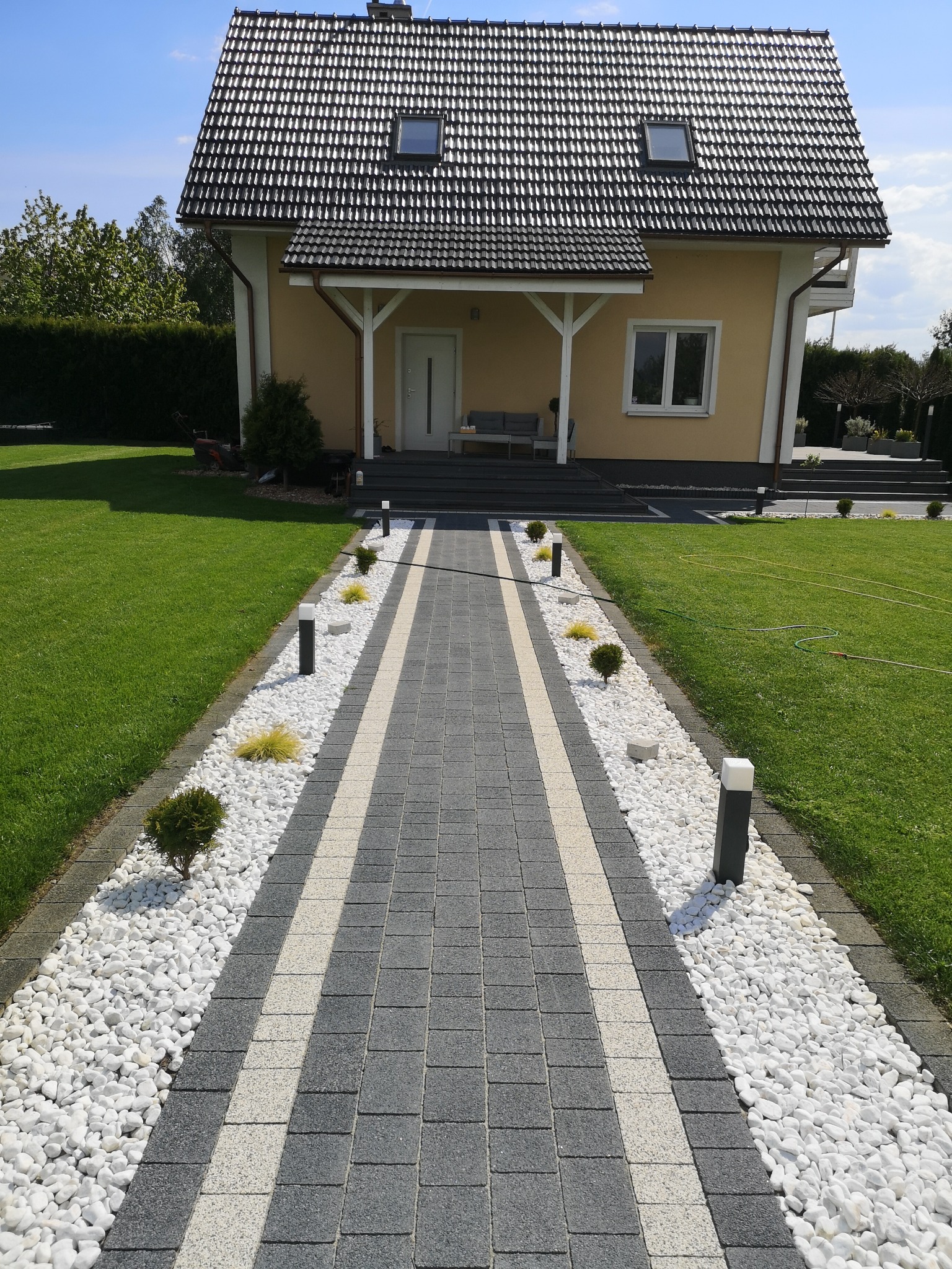 Ułożona ścieżka z kostki brukowej w odcieniach szarości i bieli, obłożona białymi kamieniami i oświetlona lampami, prowadząca do domu z zadbanym trawnikiem.