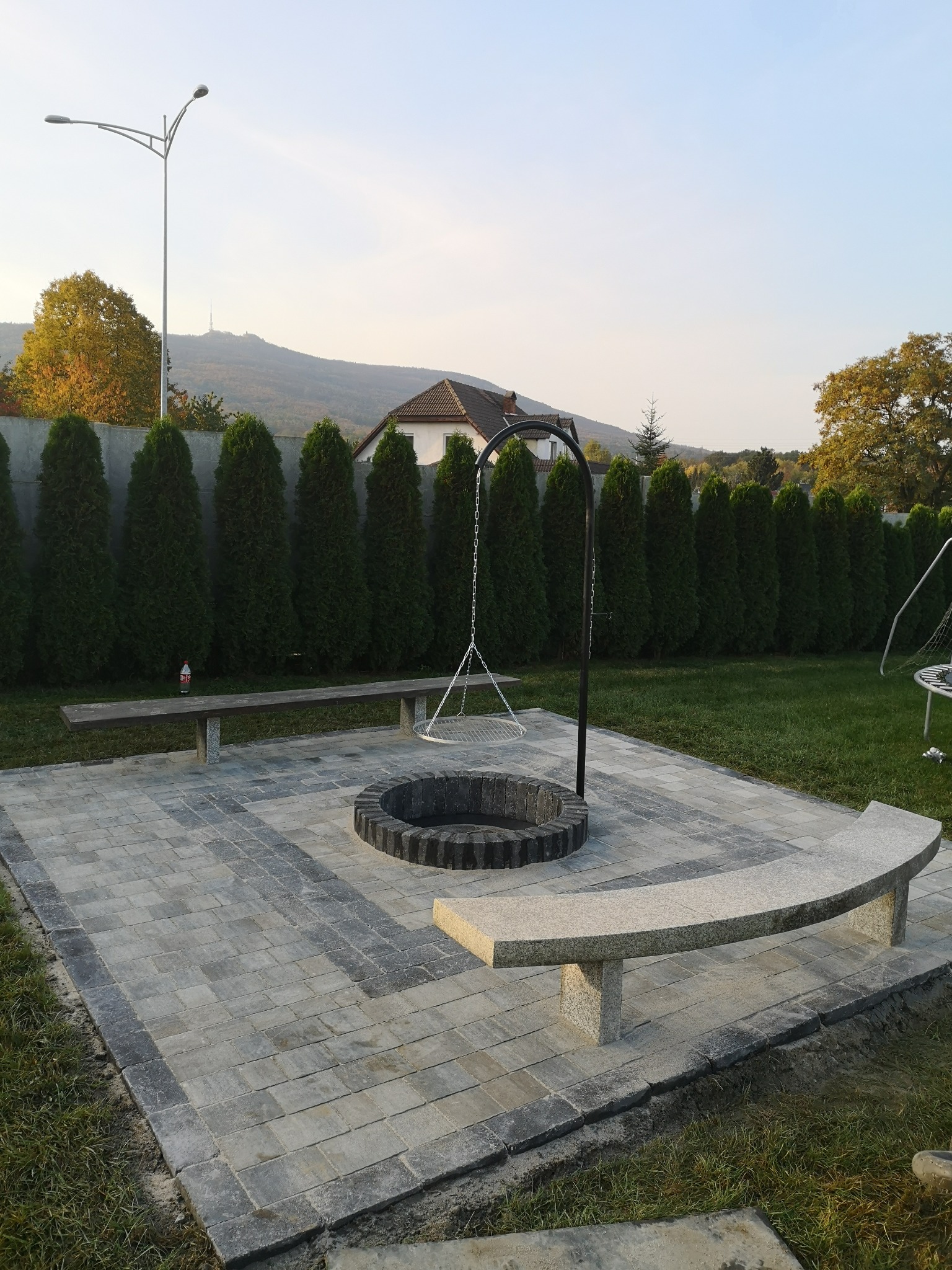 Ogród z paleniskiem otoczonym szarą kostką, ławkami z kamienia i żywopłotem z tuj, widok na górę w tle.