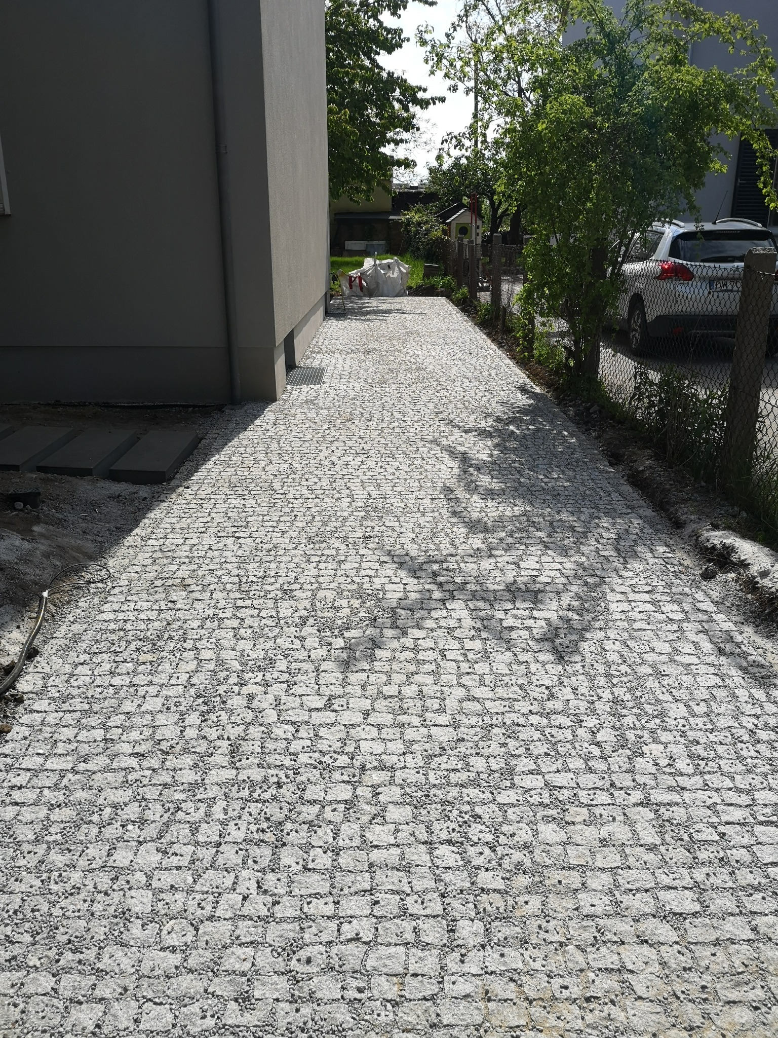 Ścieżka z jasnoszarej kostki, prowadząca wzdłuż szarego budynku, z widocznymi śladami prac ziemnych po bokach i zaparkowanym samochodem w tle.