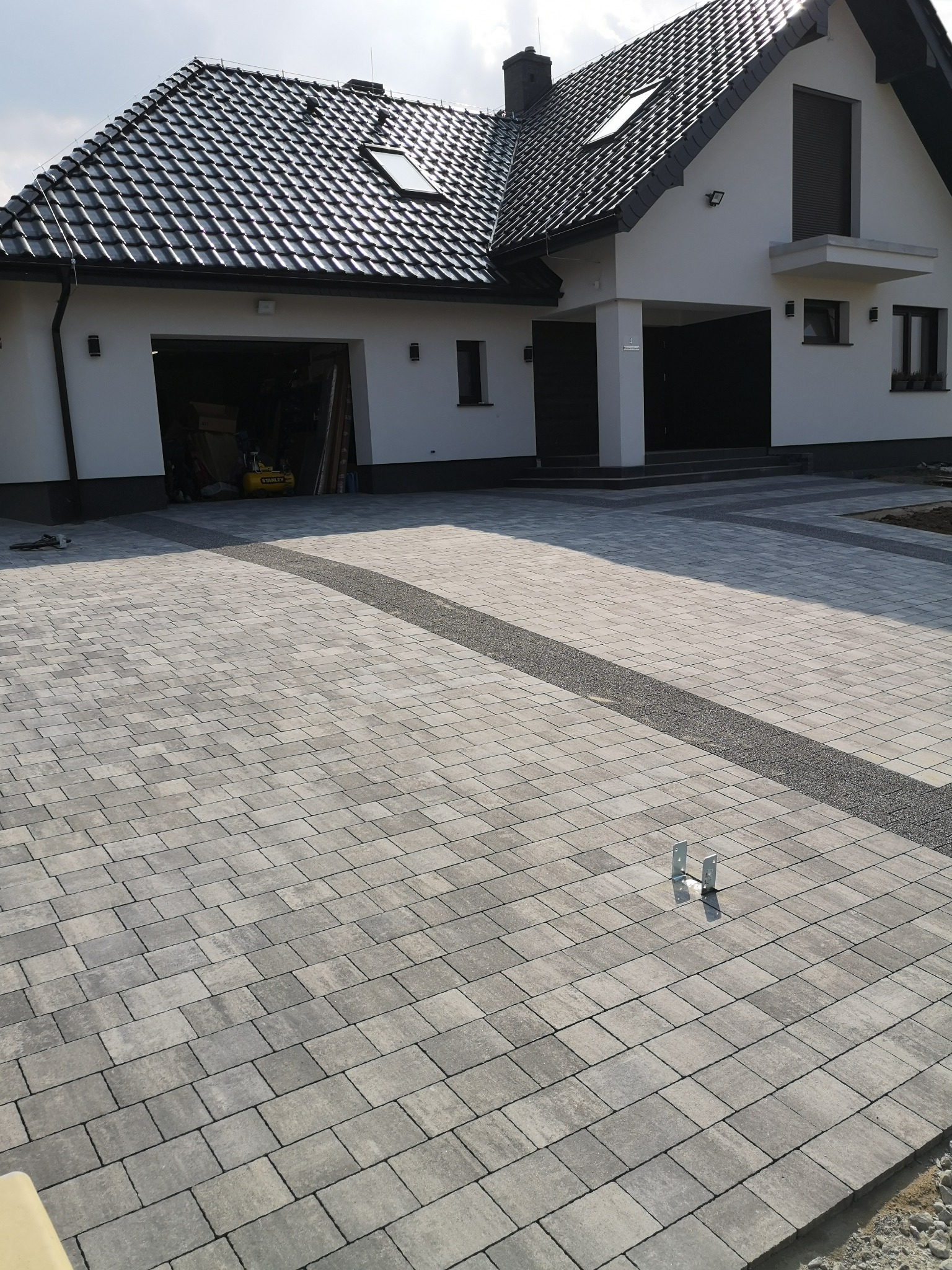 Ułożona szara kostka brukowa na podjeździe do nowoczesnego domu z garażem, z dekoracyjnym pasem z drobnego grysu.