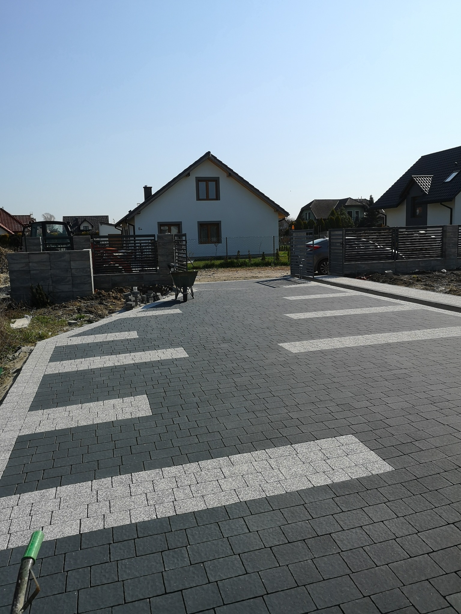 Ułożona nawierzchnia z ciemnej kostki z jasnymi wstawkami na podjeździe do domu jednorodzinnego, widoczna taczka i ogrodzenie.