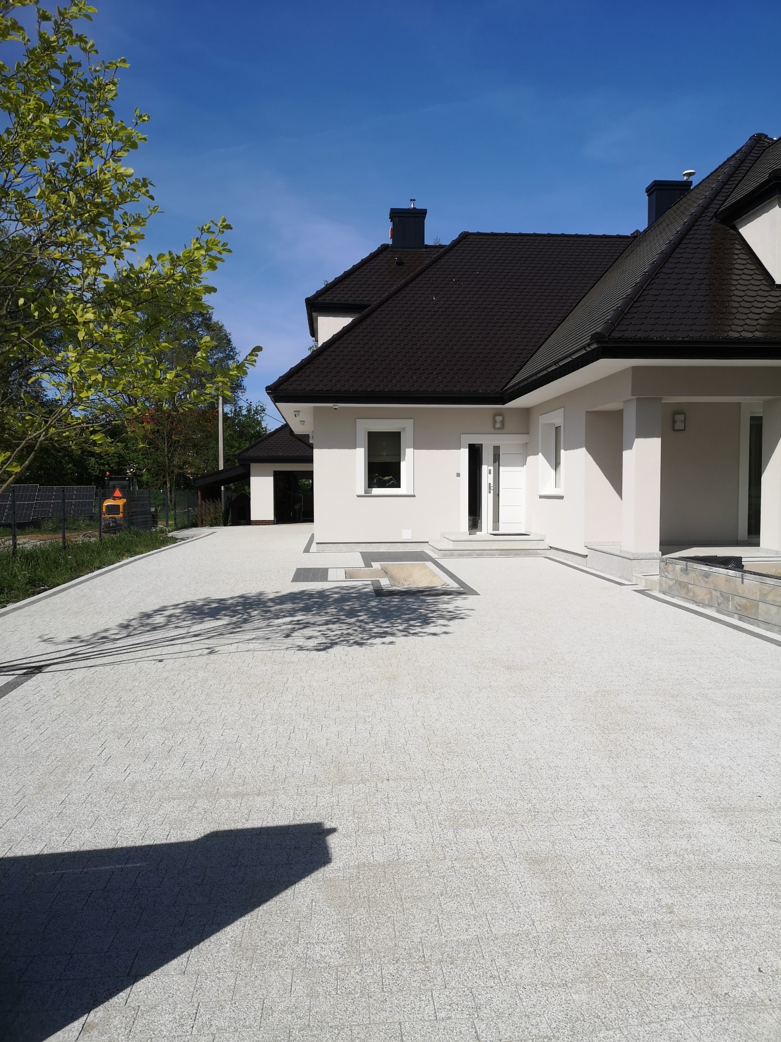 Jasnoszara kostka brukowa ułożona przed nowoczesnym domem we Wrocławiu, widoczne cienie drzew i fragment zieleni w tle.