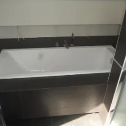 Nowoczesna biała wanna obudowana ciemną glazurą w łazience w Warszawie, widoczna częściowo pralka po lewej stronie i szklana ścianka kabiny prysznicowej po prawej.