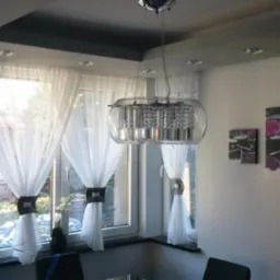 Jasne wnętrze jadalni z oknami zasłoniętymi białymi firanami, nowoczesnym żyrandolem z kryształkami i obrazami z motywem orchidei na szarej ścianie.