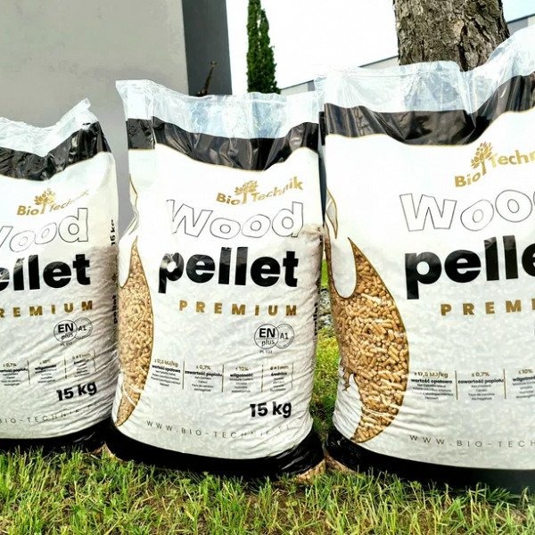 Trzy białe, foliowe worki 15 kg z napisem 'Wood pellet PREMIUM' marki Bio Technik, ustawione na trawie przed drzewem i jasną ścianą.