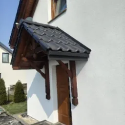 Elewacja domu z drewnianym zadaszeniem nad drzwiami wejściowymi. Biała ściana z delikatną fakturą, ciemnobrązowe elementy drewniane i szara dachówka.