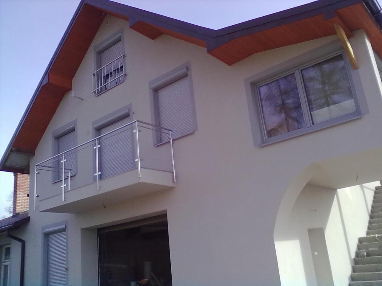 Jasna elewacja domu jednorodzinnego z balkonami i szarymi roletami okiennymi. Widoczne schody zewnętrzne i fragment garażu.