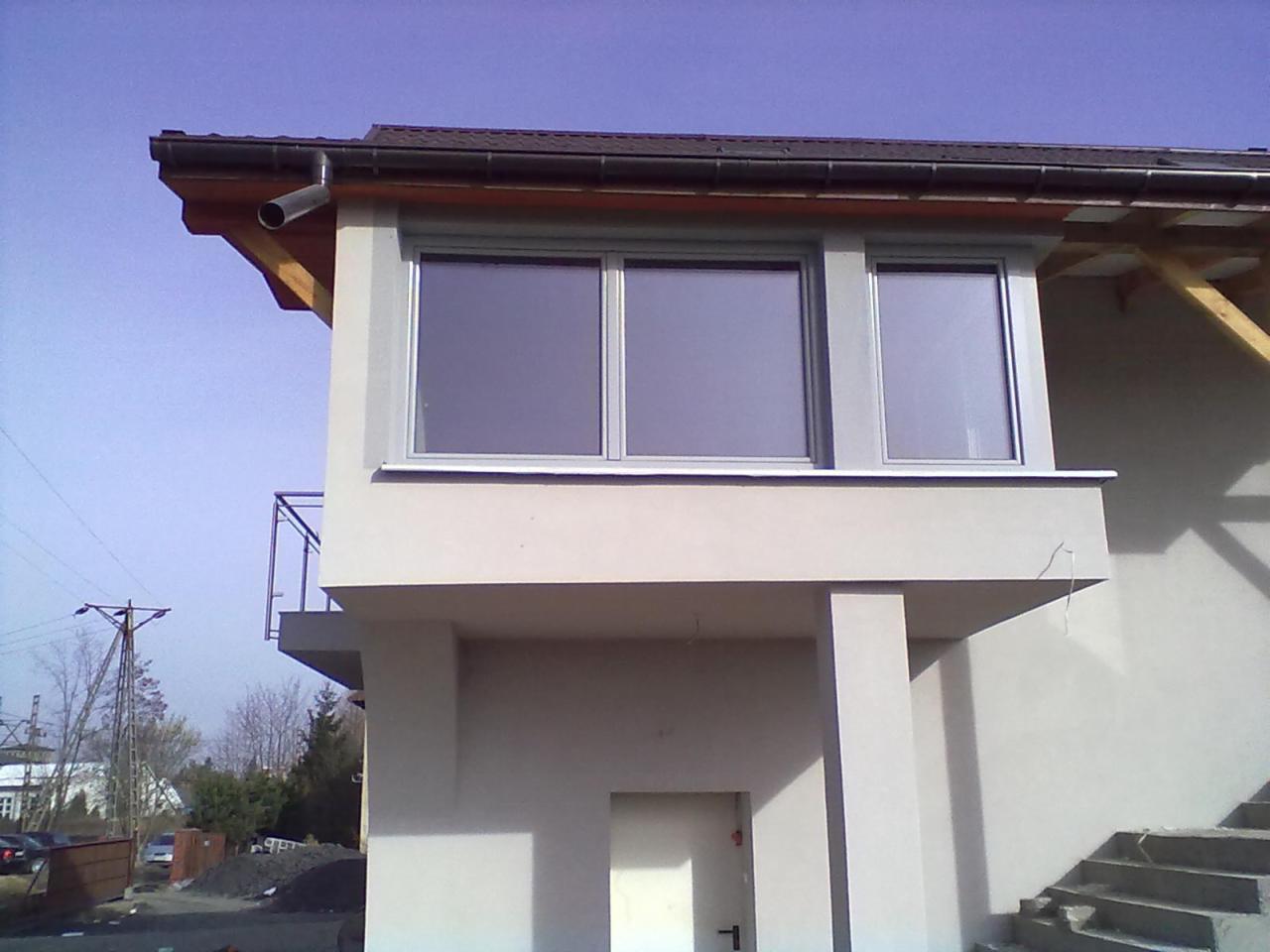 Fragment elewacji budynku z balkonem i trzema oknami w srebrnych ramach. Widoczne elementy konstrukcji dachu i rynna. Tło: błękitne niebo i drzewa.