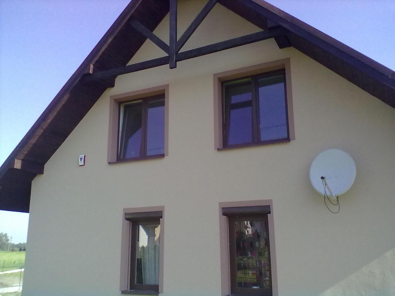 Dwukondygnacyjny dom z beżową elewacją, brązowymi ramami okiennymi i satelitą. Widoczne drewniane elementy dekoracyjne pod dachem.