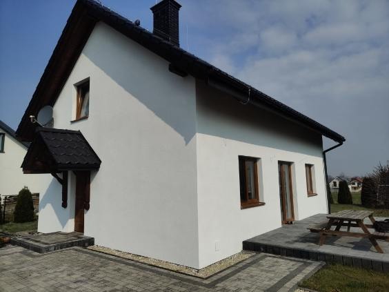 Biała elewacja domu jednorodzinnego z ciemnymi oknami i dachem, widok z boku, kostka brukowa na podjeździe, stół piknikowy na tarasie, słoneczny dzień.