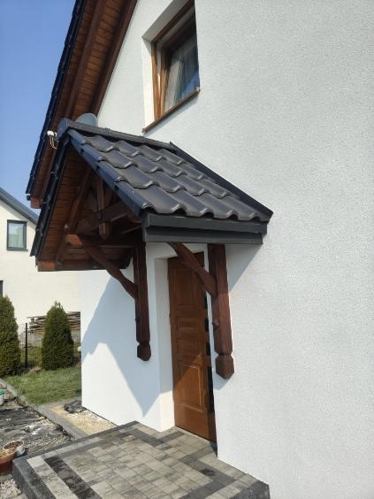 Elewacja domu z drewnianym zadaszeniem nad drzwiami wejściowymi. Biała ściana z delikatną fakturą, ciemnobrązowe elementy drewniane i szara dachówka.