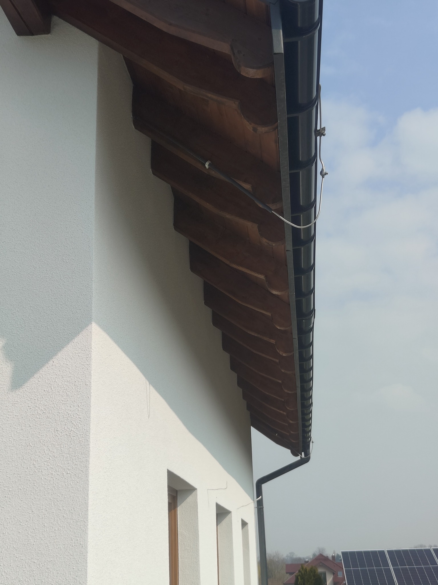 Elewacja budynku z drewnianym podbitkiem i czarnymi rynnami, widok z dołu, z fragmentem paneli słonecznych w tle. Teksturowana, biała ściana.