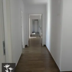 Długi, jasny korytarz z drewnopodobną podłogą, białymi ścianami i kilkoma parami białych drzwi, widok w głąb mieszkania z perspektywy logo firmy projektowej w lewym dolnym rogu.