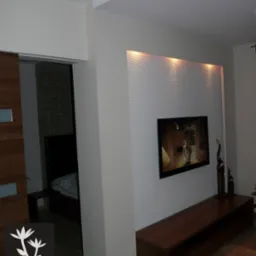 Salon z telewizorem na ścianie z poziomymi żłobieniami, podświetlonymi od góry, drewnianą szafką RTV, widok na sypialnię przez otwarte drzwi, logo firmy projektującej wnętrza w lewym dolnym rogu.