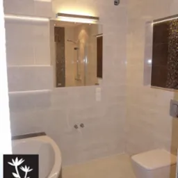 Jasna łazienka z beżowymi płytkami, mozaiką na obudowie wanny i w ramie lustra, podświetlane lustro, wisząca toaleta, logo firmy w lewym dolnym rogu.