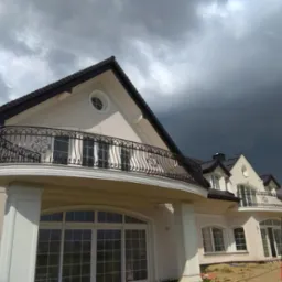 Elewacja domu z balkonami z ozdobnymi, kutymi balustradami, widok z dołu na tle ciemnego, burzowego nieba. W prawym dolnym rogu widoczne logo firmy projektowej.