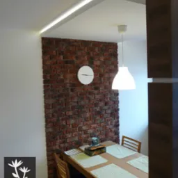 Jadalnia z ceglaną ścianą akcentową, oświetleniem LED w suficie podwieszanym, minimalistycznym zegarem i drewnianym stołem nakrytym dla czterech osób.