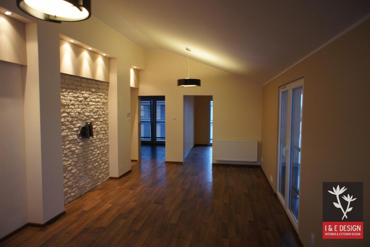 Jasne wnętrze po remoncie: drewniana podłoga, ściana z dekoracyjnej cegły i nowoczesne oświetlenie. Widoczne okna i drzwi z żaluzjami. Minimalistyczny design.