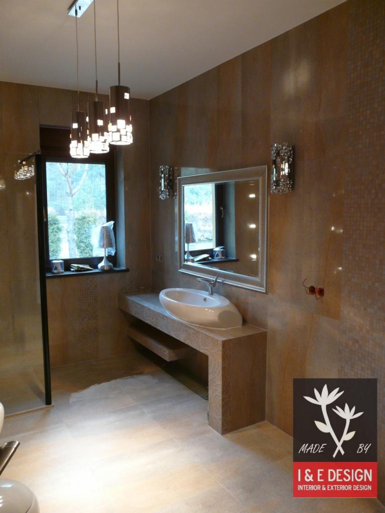 Jasna łazienka z beżowymi płytkami, umywalką nablatową i wiszącymi lampami. Widoczny fragment kabiny prysznicowej i okno z widokiem na zieleń.