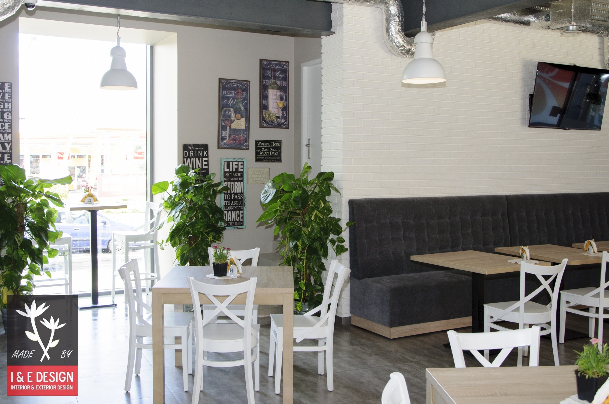 Jasne wnętrze kawiarni lub restauracji z białymi krzesłami, drewnianymi stołami, szarą tapicerowaną ławą pod ścianą z białej cegły oraz dekoracyjnymi roślinami doniczkowymi i plakatami na ścianach.