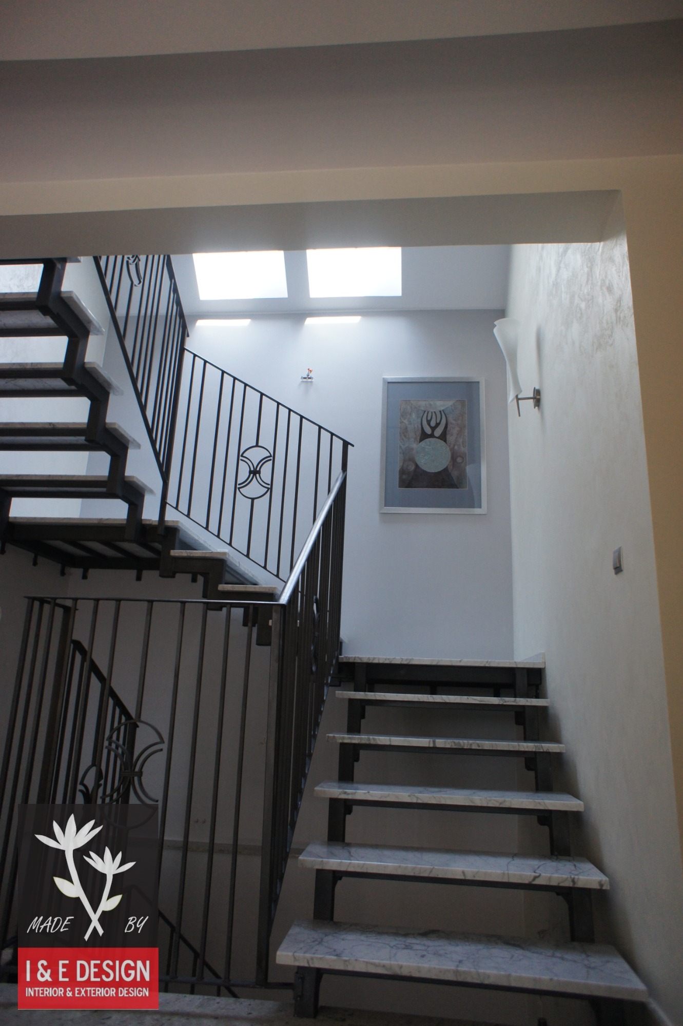 Klatka schodowa z marmurowymi stopniami i metalową balustradą z ozdobnym motywem, oświetlona światłem dziennym z okien dachowych i lampą ścienną, z obrazem na ścianie i logo firmy I & E Design.