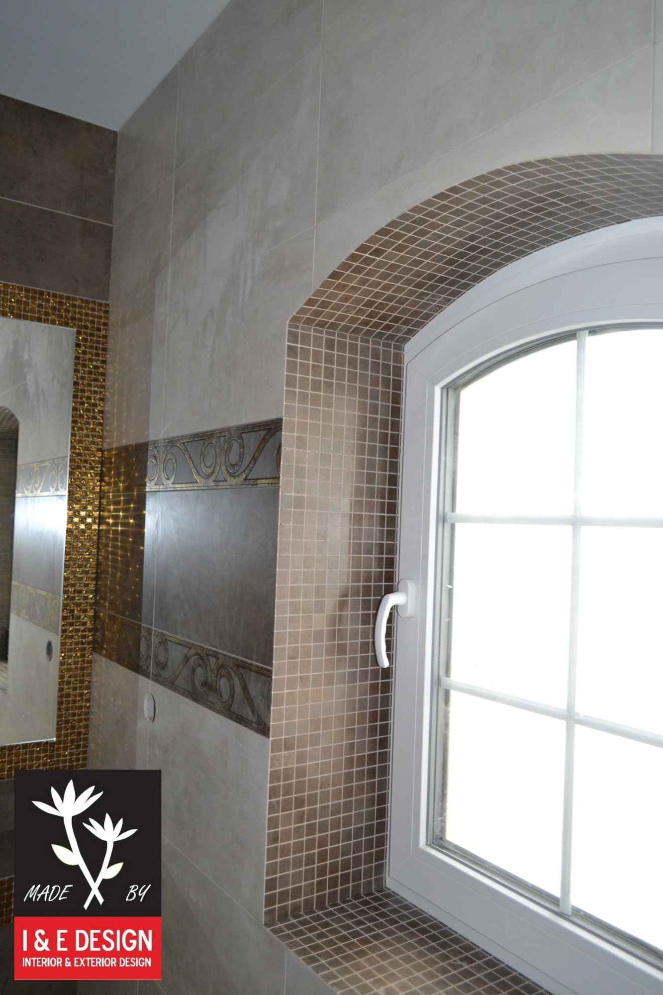 Wykończona łazienka z beżowymi płytkami i mozaiką, okno łukowe z białymi ramami, widoczne logo firmy w rogu.