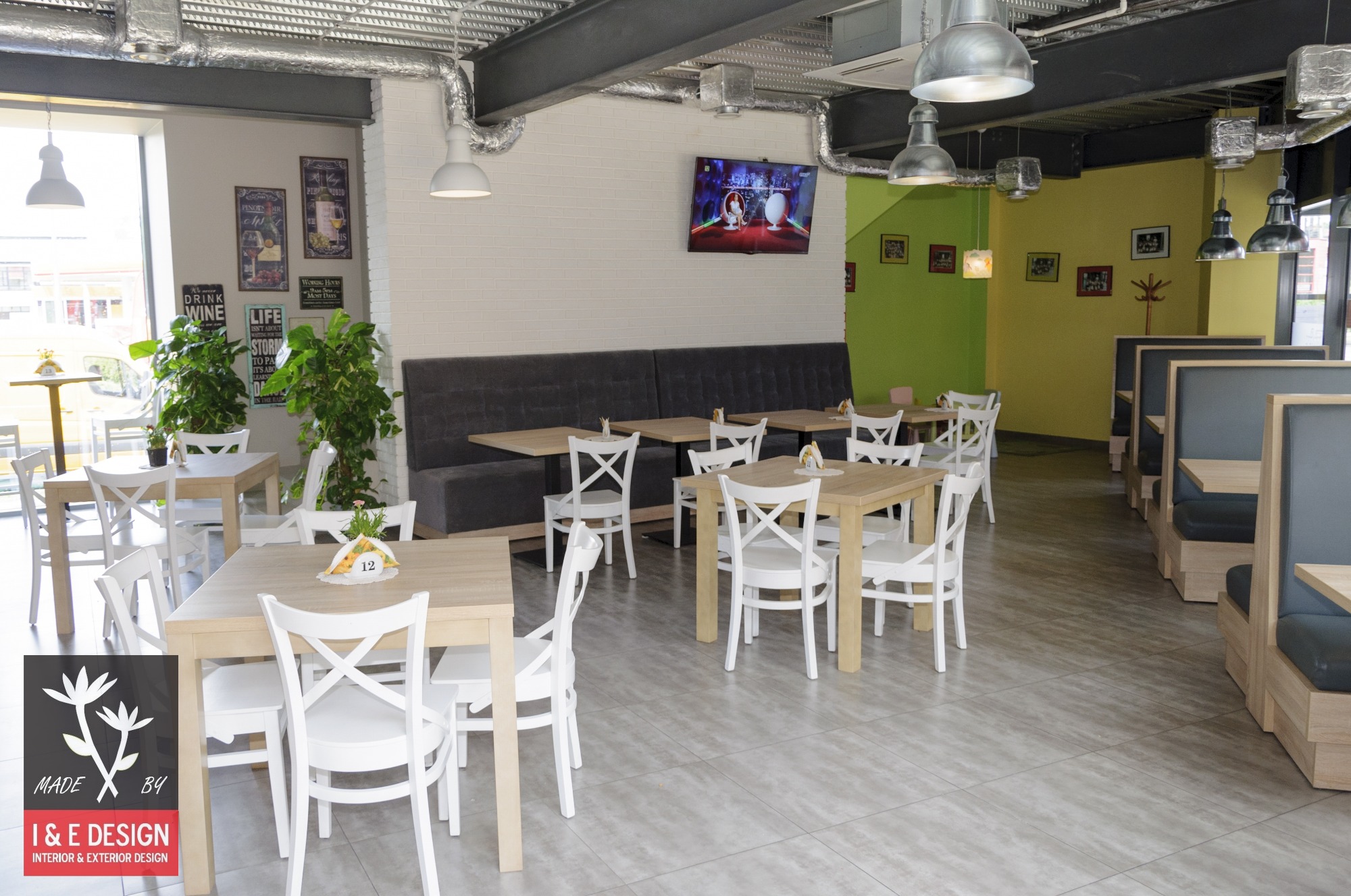 Jasne wnętrze restauracji z drewnianymi stołami i białymi krzesłami, szarą podłużną kanapą, płytkami podłogowymi imitującymi kamień, białymi ceglanymi ścianami, elementami wentylacji pod sufitem...