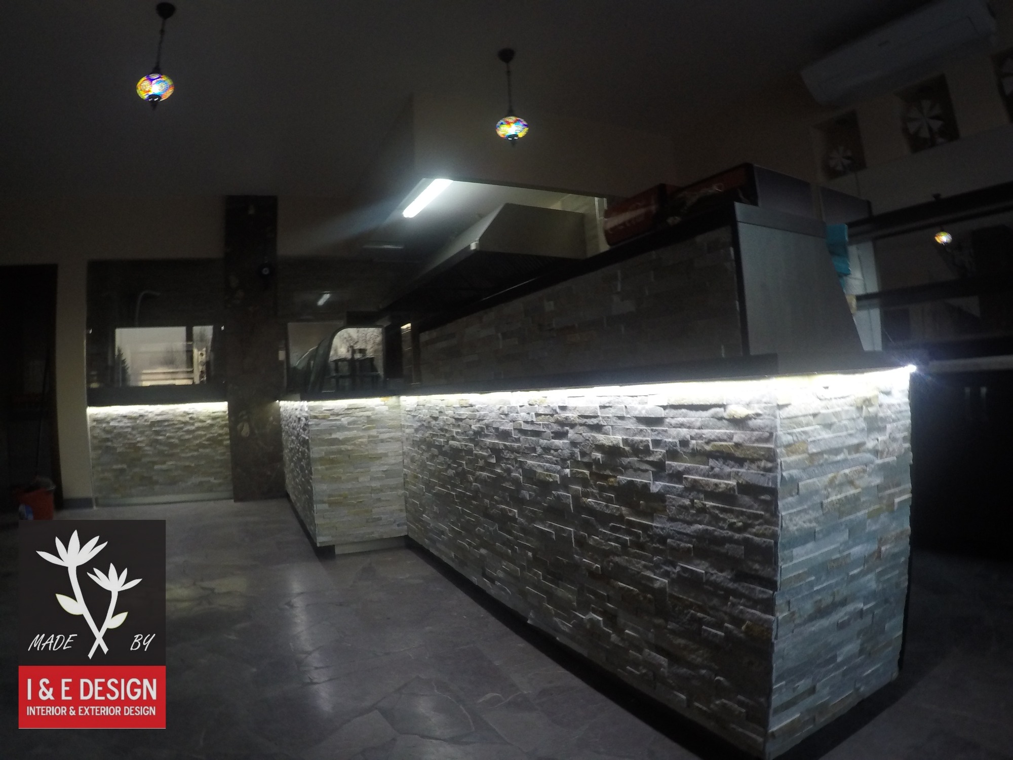 Wykończona ściana z kamienia dekoracyjnego z podświetleniem LED, widoczna w przestrzeni komercyjnej, z logo firmy projektowej w lewym dolnym rogu.