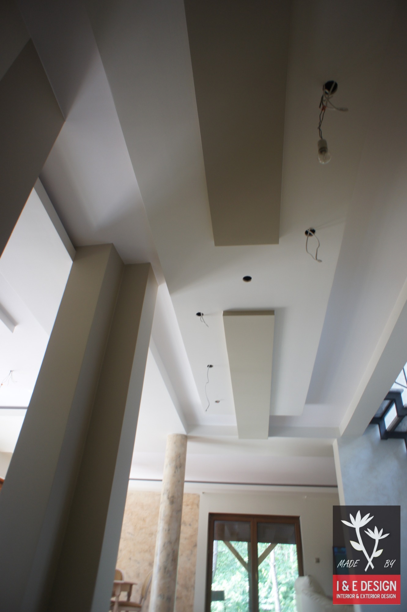 Wykończony sufit z podwieszanymi elementami i punktami oświetleniowymi z widocznymi przewodami, w tle kolumna i okno z widokiem na zieleń.