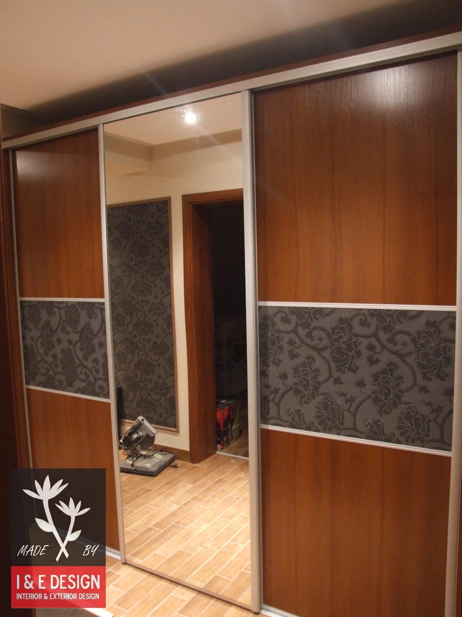 Szafa przesuwna z drewnianymi panelami i lustrami, ozdobiona szarym wzorem kwiatowym; widoczne narzędzie na podłodze i logo firmy w rogu.