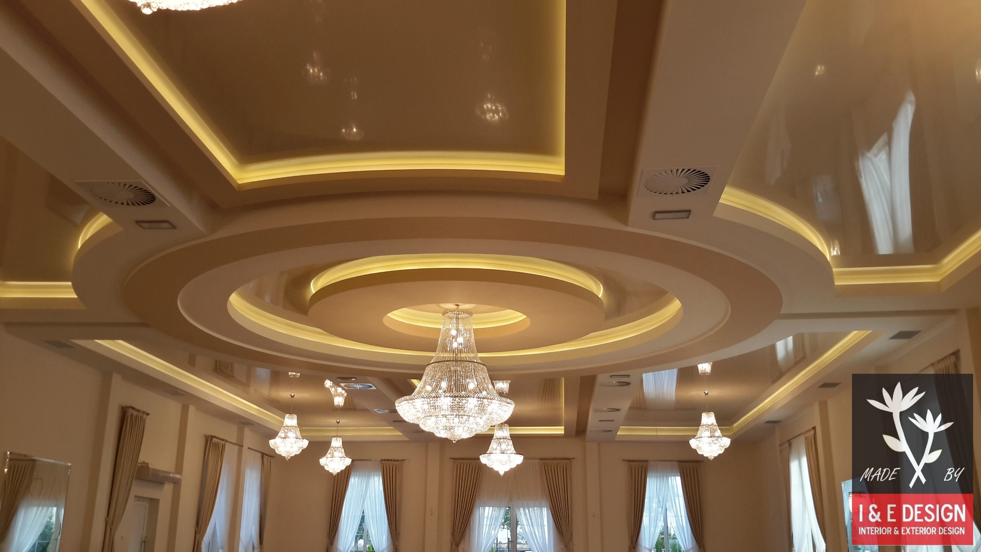 Ozdobny sufit podwieszany z połyskiem, wielopoziomowy, z oświetleniem LED i centralnym żyrandolem kryształowym w jasnym wnętrzu sali balowej.