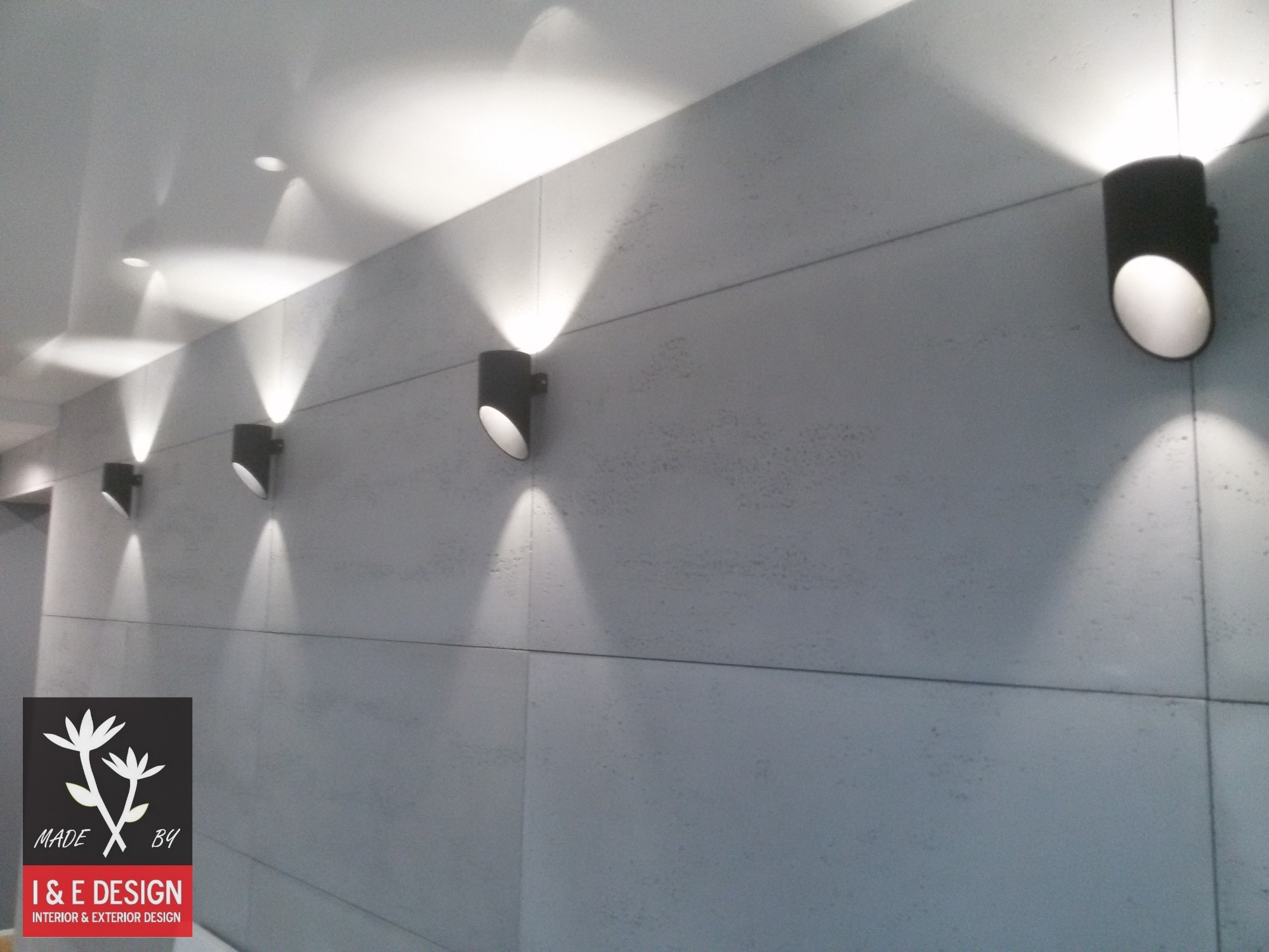 Szara ściana z widocznymi spoinami między płytami, oświetlona szeregiem czarnych, cylindrycznych kinkietów dających stożkowe światło oraz logo firmy I & E Design w lewym dolnym rogu.