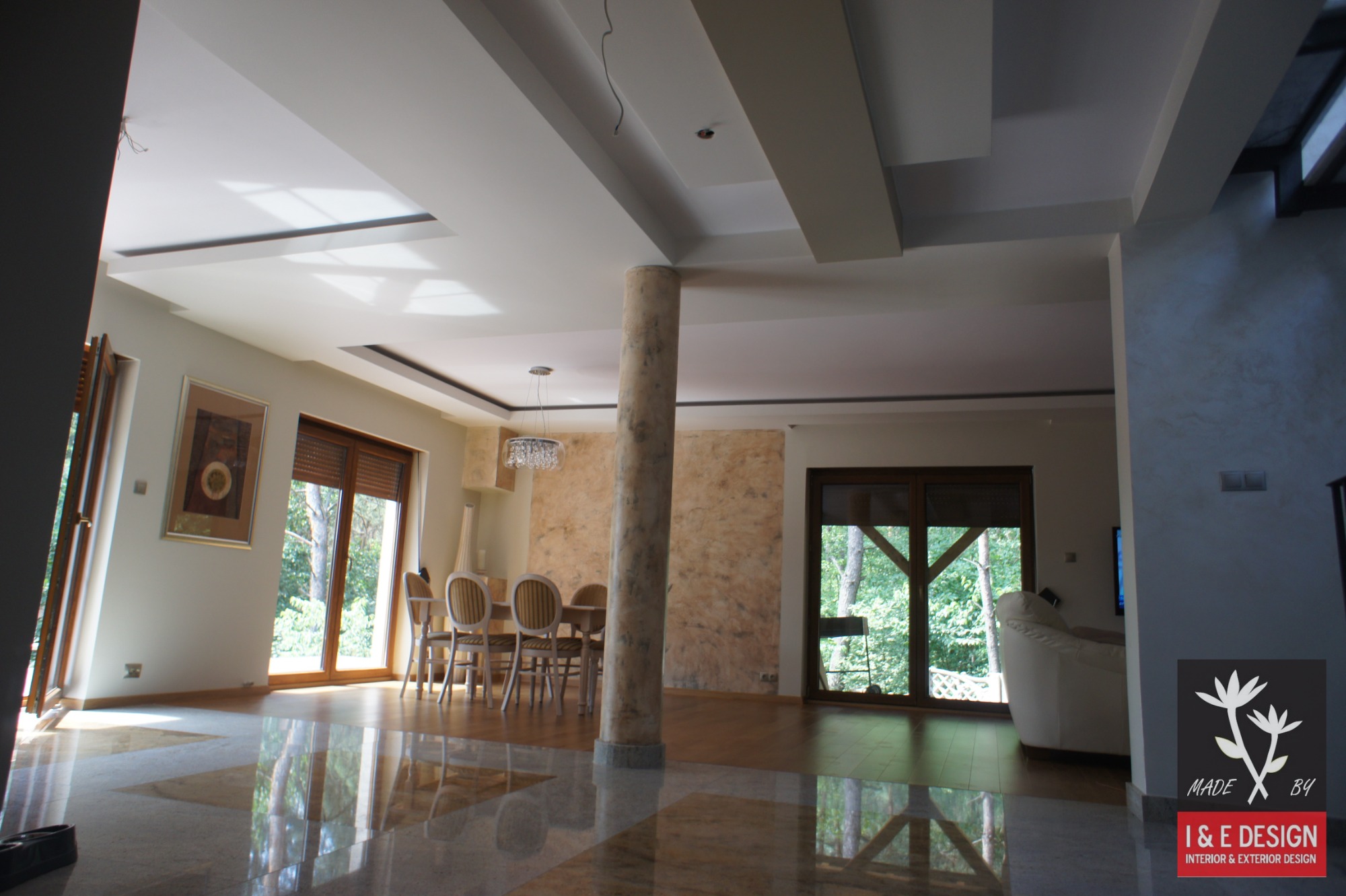 Przestronne wnętrze z marmurową podłogą, kolumną dekoracyjną i ścianą z efektem trawertynu, widok na ogród przez duże okna.