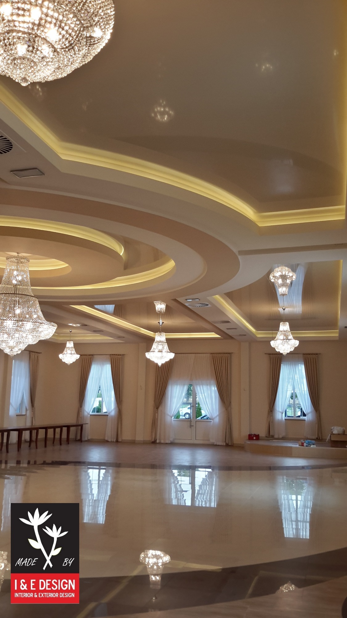 Eleganckie wnętrze sali balowej z marmurową podłogą, ozdobnym sufitem podwieszanym z oświetleniem LED i kryształowymi żyrandolami, widoczne okna z jasnymi zasłonami.