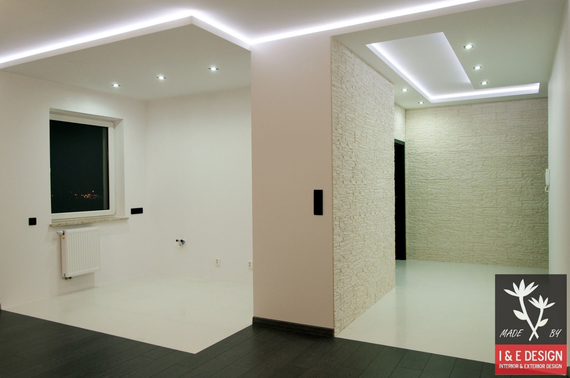 Nowoczesne wnętrze z białymi ścianami, podwieszanym sufitem z oświetleniem LED, fragmentem ściany wykończonej dekoracyjną okładziną imitującą kamień i kontrastującą, ciemną podłogą w strefie wejścia.