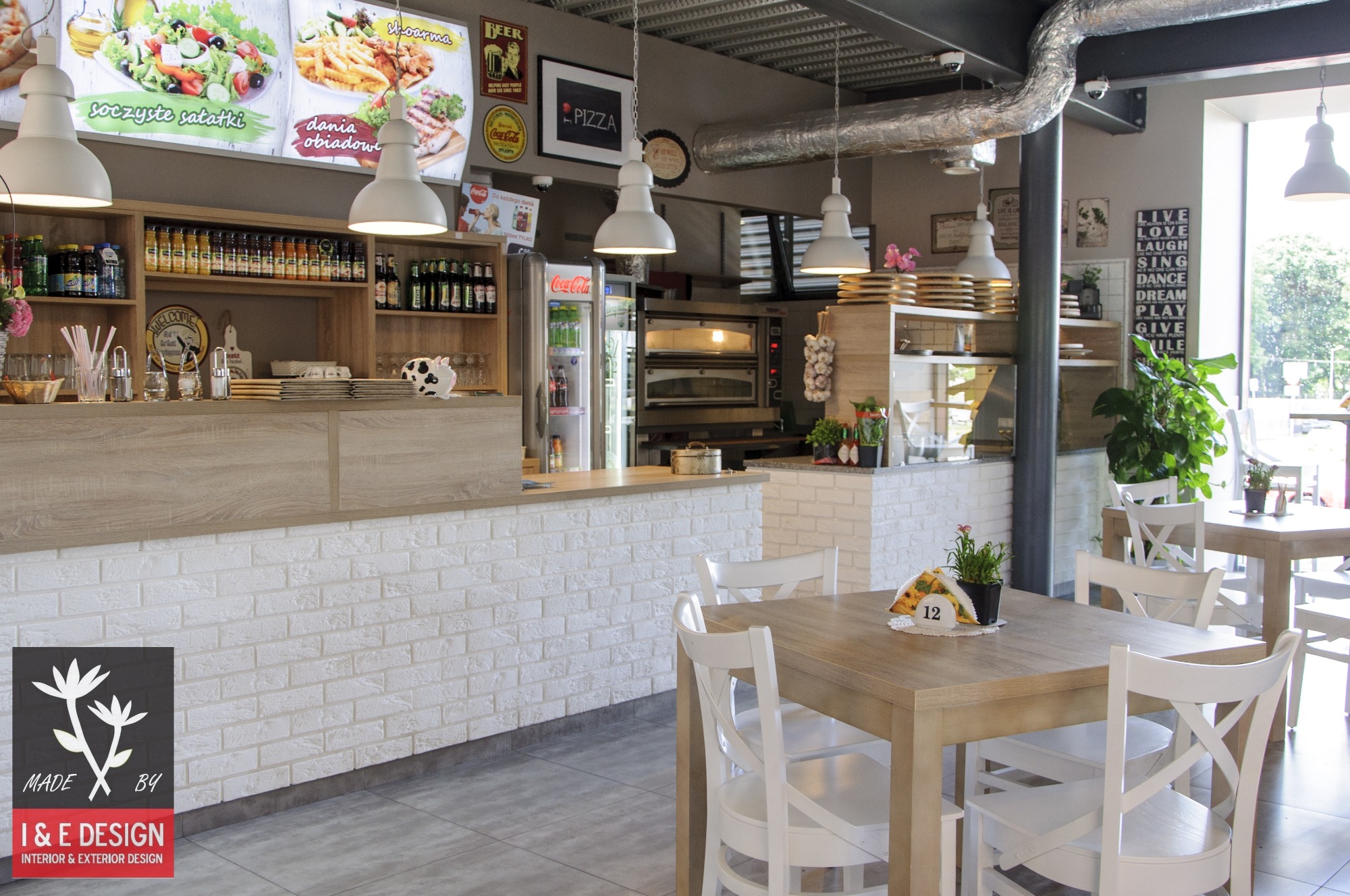 Wnętrze restauracji z białymi ceglanymi ścianami, drewnianymi blatami, białymi krzesłami i wiszącymi lampami, widok na zaplecze z piecem do pizzy i lodówką Coca-Cola.