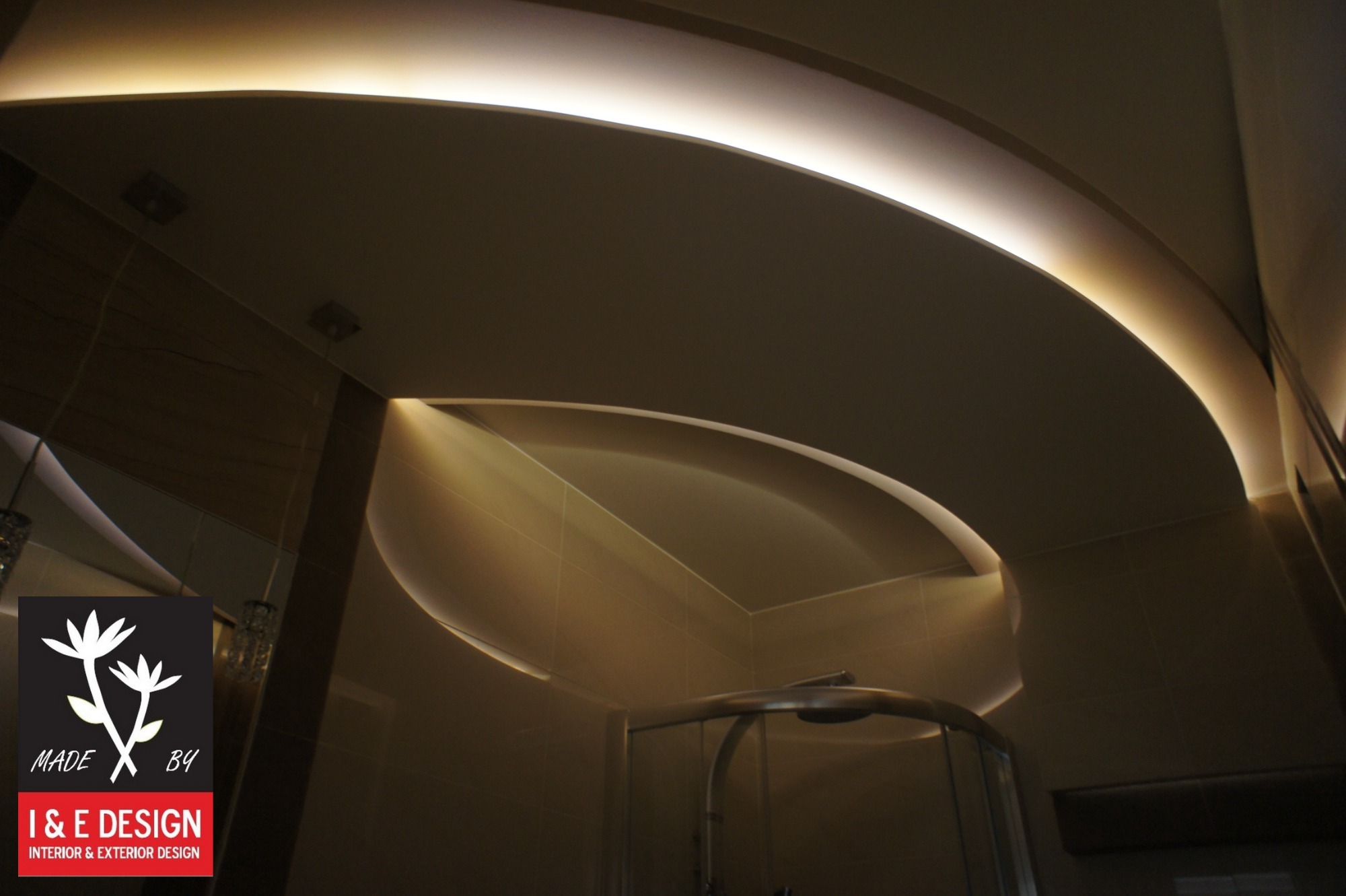 Łazienka z podwieszanym, podświetlanym sufitem o zaokrąglonych kształtach i kabiną prysznicową z chromowanymi elementami