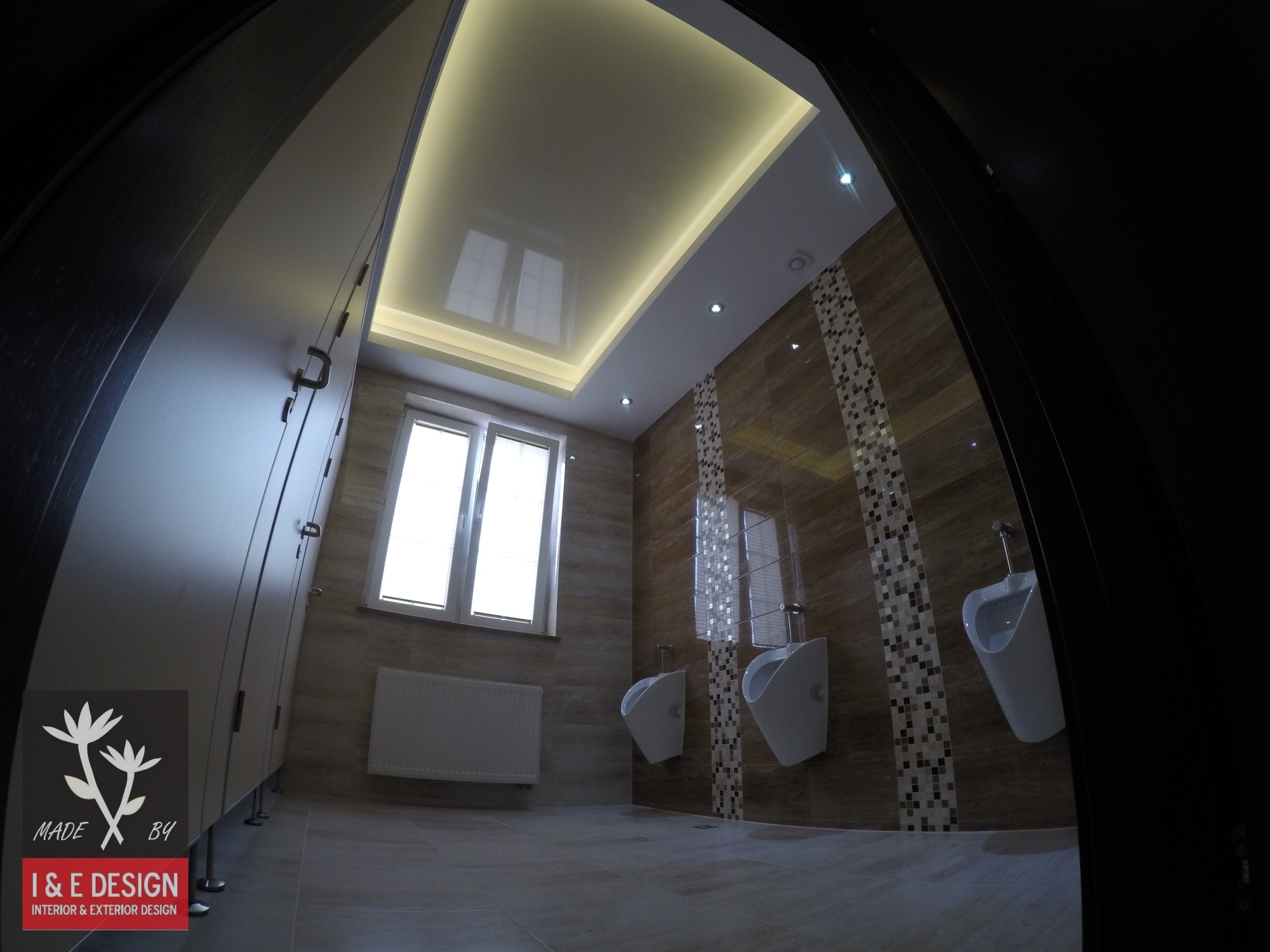 Nowoczesna toaleta publiczna z trzema pisuarami, brązowymi płytkami z mozaikowymi wstawkami, podświetlanym sufitem napinanym z oknem i białym grzejnikiem pod oknem, widziana zza częściowo otwartych...