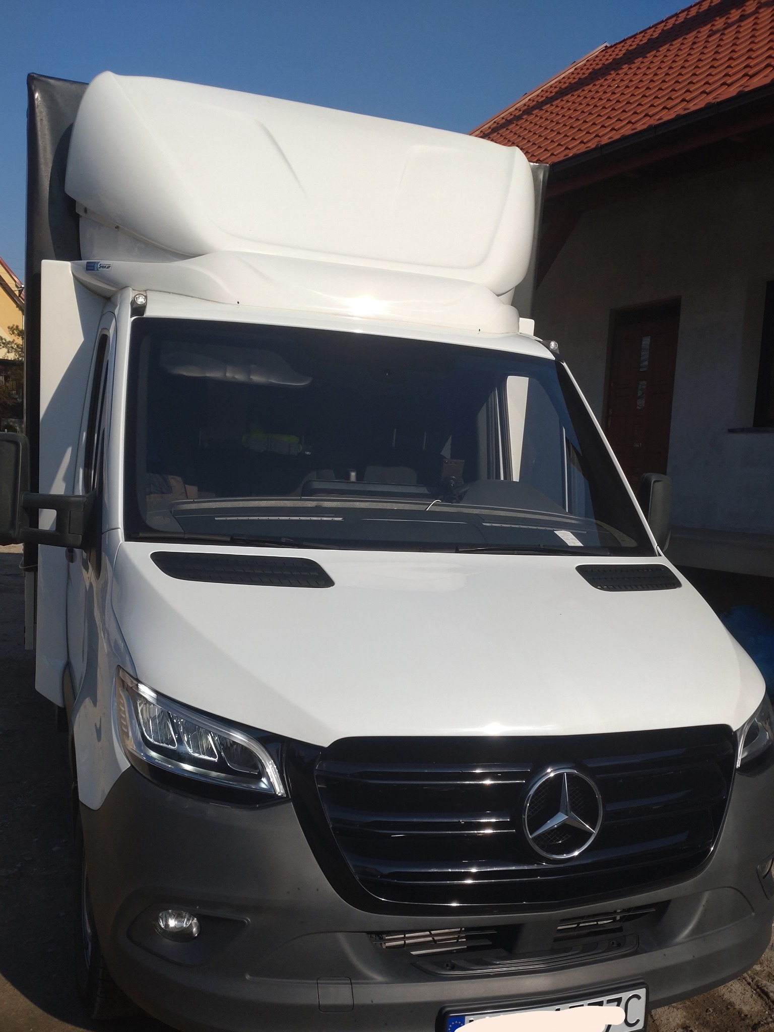 Biały samochód dostawczy Mercedes-Benz z zabudową kontenerową na tle domu z czerwoną dachówką, widok z przodu.